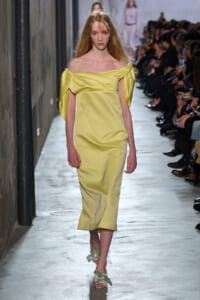 Model on a runway wearing a pale yellow off-the-shoulder midi dress with sculpted draped sleeves and matching bow-strap sandals.