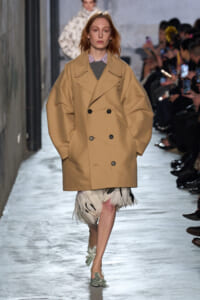 Model walks a runway in a tan oversized double-breasted trench coat with a feathered cream skirt and mint-green flats, audience in background