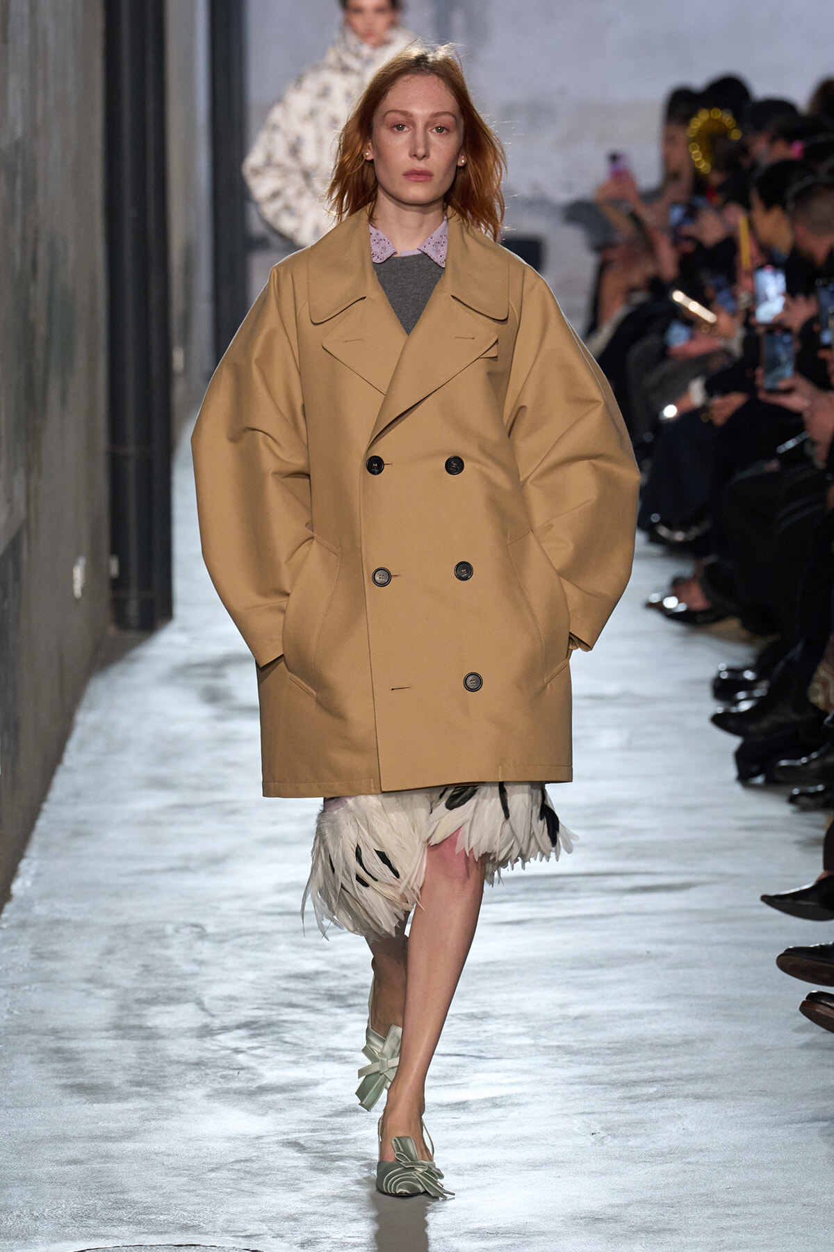 Model walks a runway in a tan oversized double-breasted trench coat with a feathered cream skirt and mint-green flats, audience in background