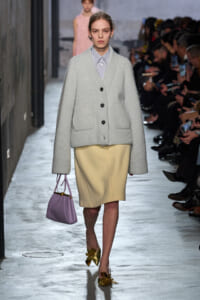 Runway model in a light gray oversized cardigan with patch pockets, paired with a beige knee-length skirt and gold bow sandals, carrying a lilac handbag.
