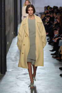 Model walking runway in a pale yellow oversized coat over a sage-green dress, amid a fashion show audience on both sides.