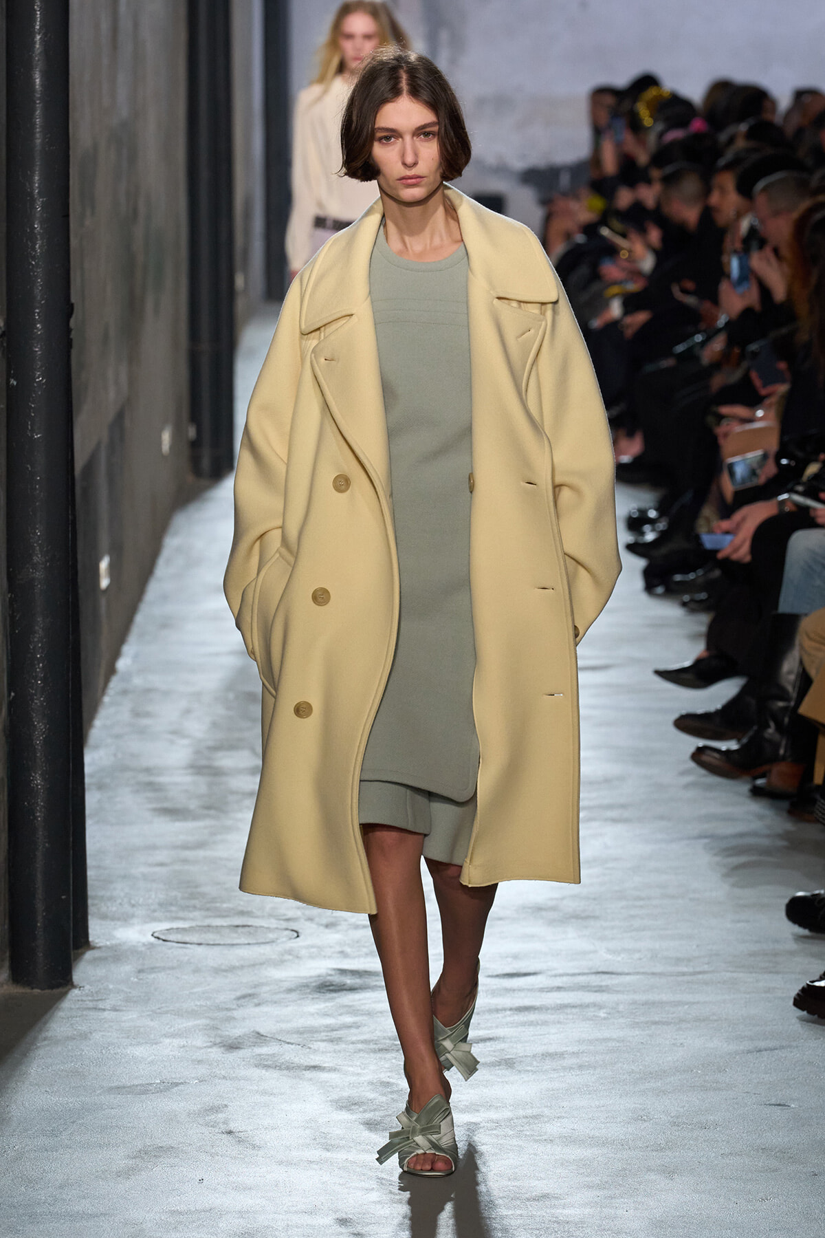 Model walking runway in a pale yellow oversized coat over a sage-green dress, amid a fashion show audience on both sides.