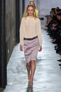 Models walks a runway wearing a cream blouse with pleated front and a lilac pencil skirt with a studded black belt.