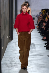 Runway model wearing a bright red turtleneck sweater and brown corduroy wide-leg pants walking the catwalk with audience in the background, hands in pockets.
