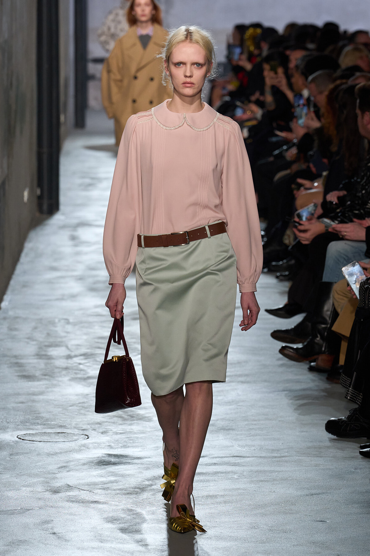 Runway model in a pale pink blouse with a lace collar and a beige pencil skirt, brown belt, yellow bow-embellished heels, and a burgundy handbag.