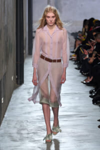 Model walking a runway in a sheer pink button-down dress with a brown belt and pale green sandals; audience seated along the catwalk to the right.