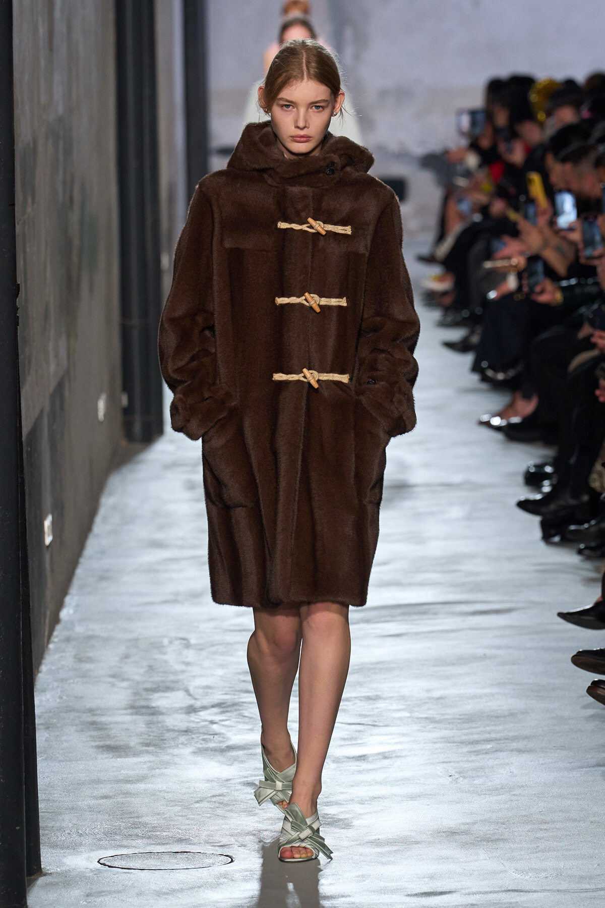 Model on runway wearing a knee-length brown fur coat with beige rope toggle closures, audience visible in the background.
