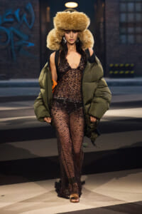 Model walking the runway in a sheer black lace jumpsuit, oversized olive puffer coat, and a voluminous fur hat.