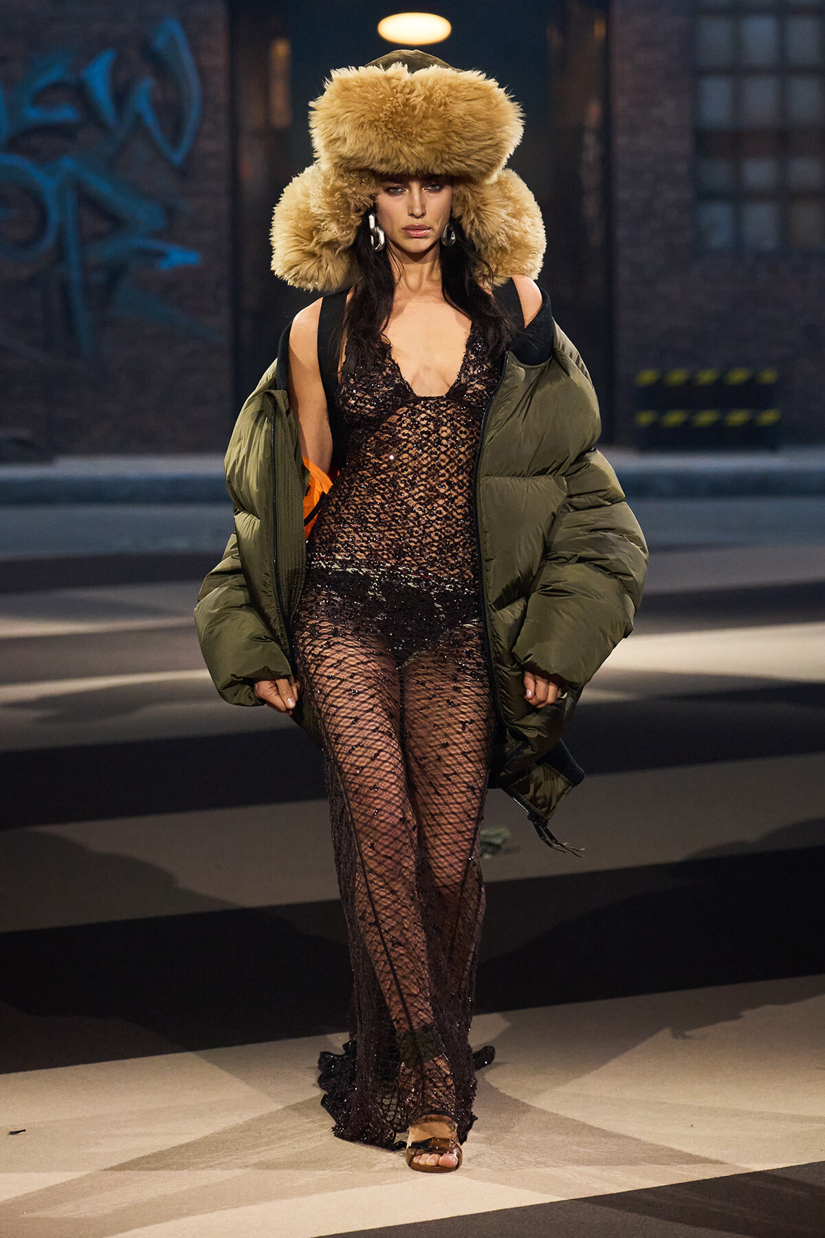 Model walking the runway in a sheer black lace jumpsuit, oversized olive puffer coat, and a voluminous fur hat.