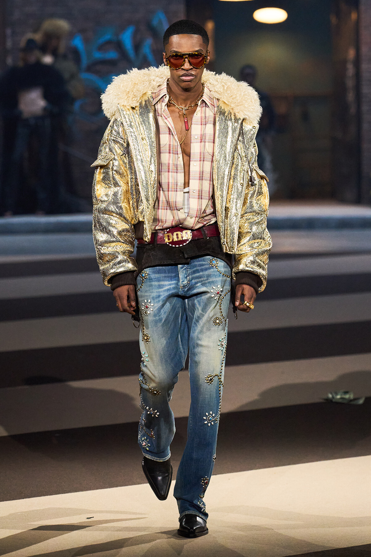 Male model wearing a shiny gold bomber with a fur-lined collar, plaid shirt, embellished jeans, and sunglasses on a runway venue