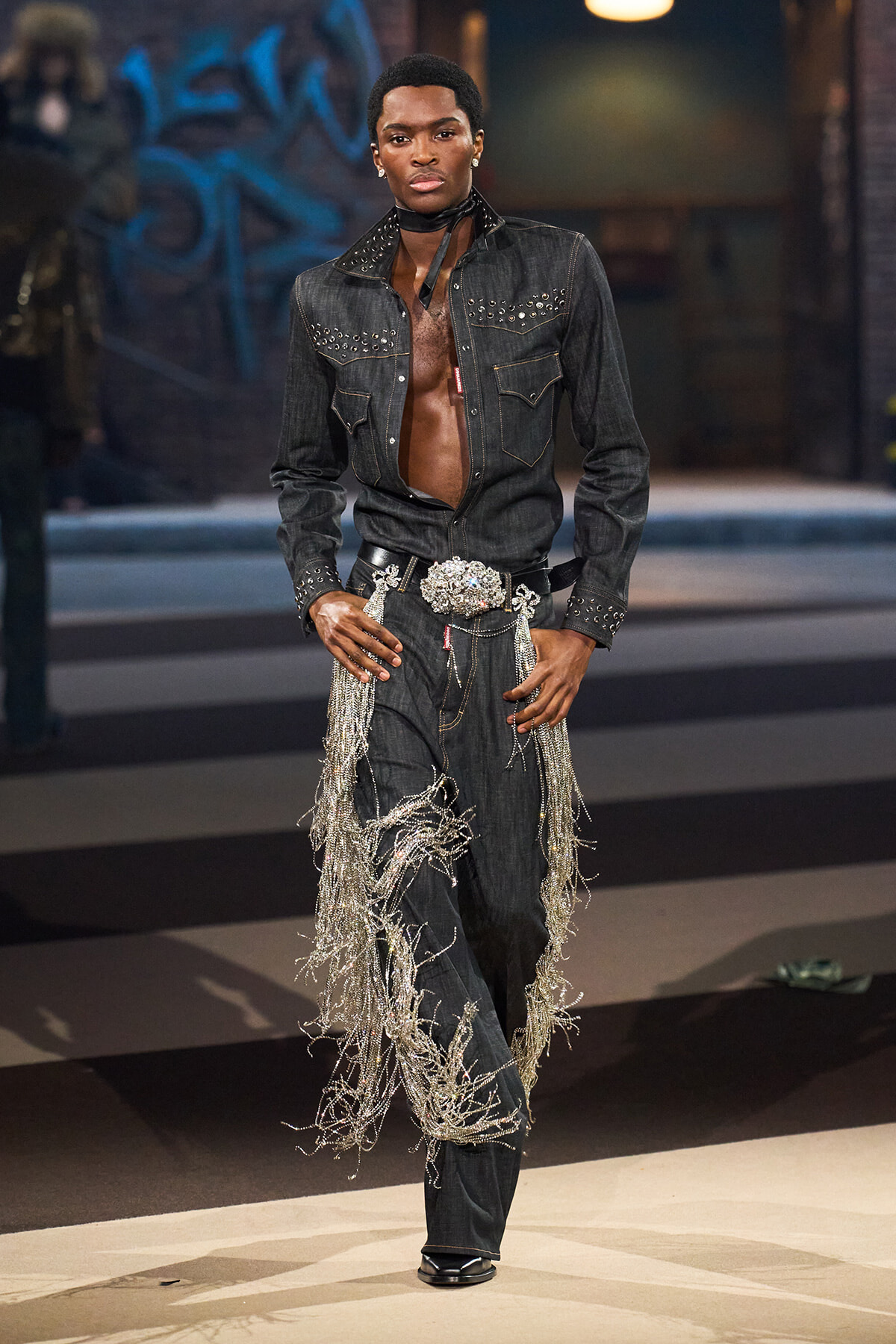 Male model on a runway wearing a dark denim denim-on-denim look with studded jacket, open chest, and long silver fringe chains on the pants belt area.