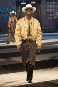 Runway model wearing a cream cowboy hat, gold bomber jacket with fur trim, mesh top, and oversized brown cargo pants with boots.