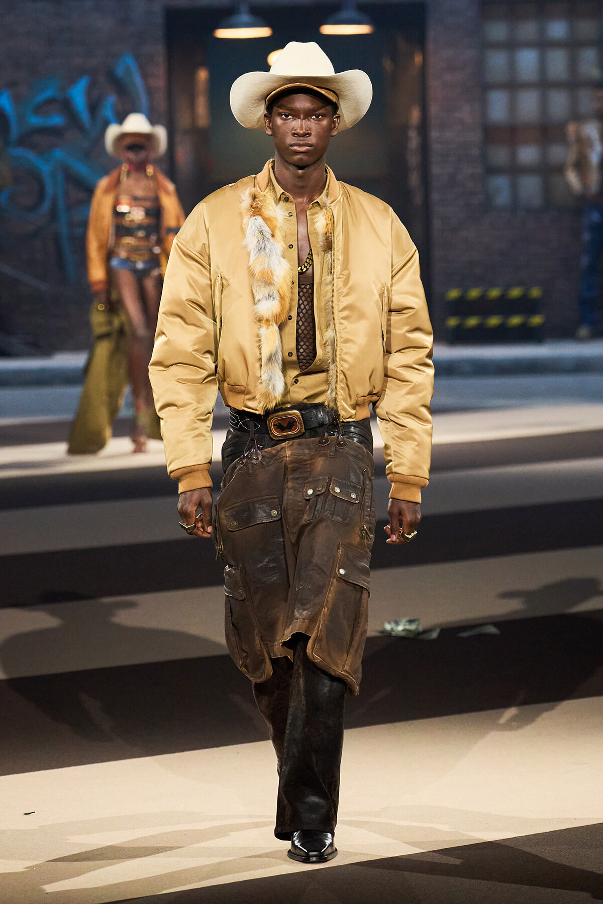 Runway model wearing a cream cowboy hat, gold bomber jacket with fur trim, mesh top, and oversized brown cargo pants with boots.
