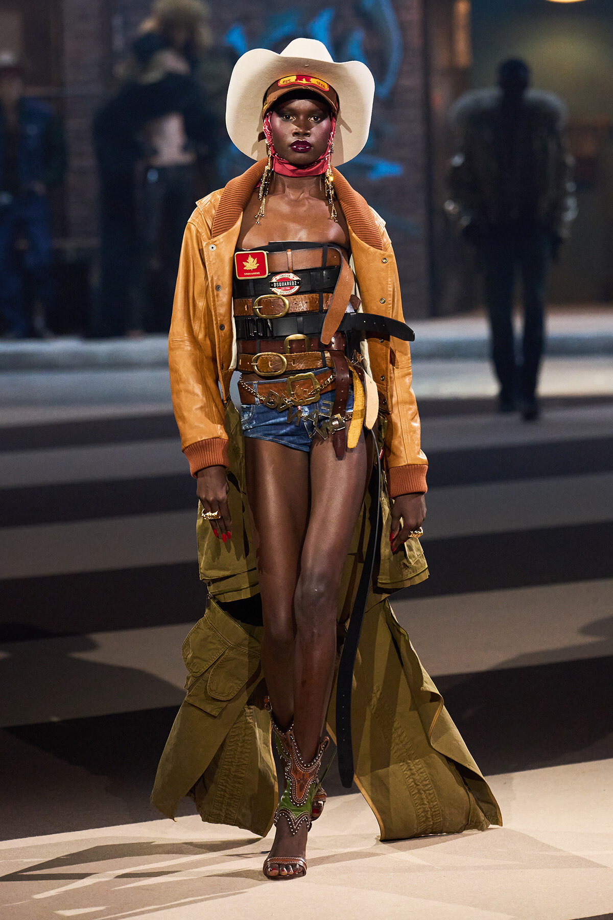 Model wearing a white cowboy hat, tan trench coat, and stacked brown belts over denim shorts on a runway