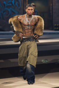 Tattooed male model wearing a fur coat draped over his shoulders, cargo shorts, and jeans, posing in an urban setting.