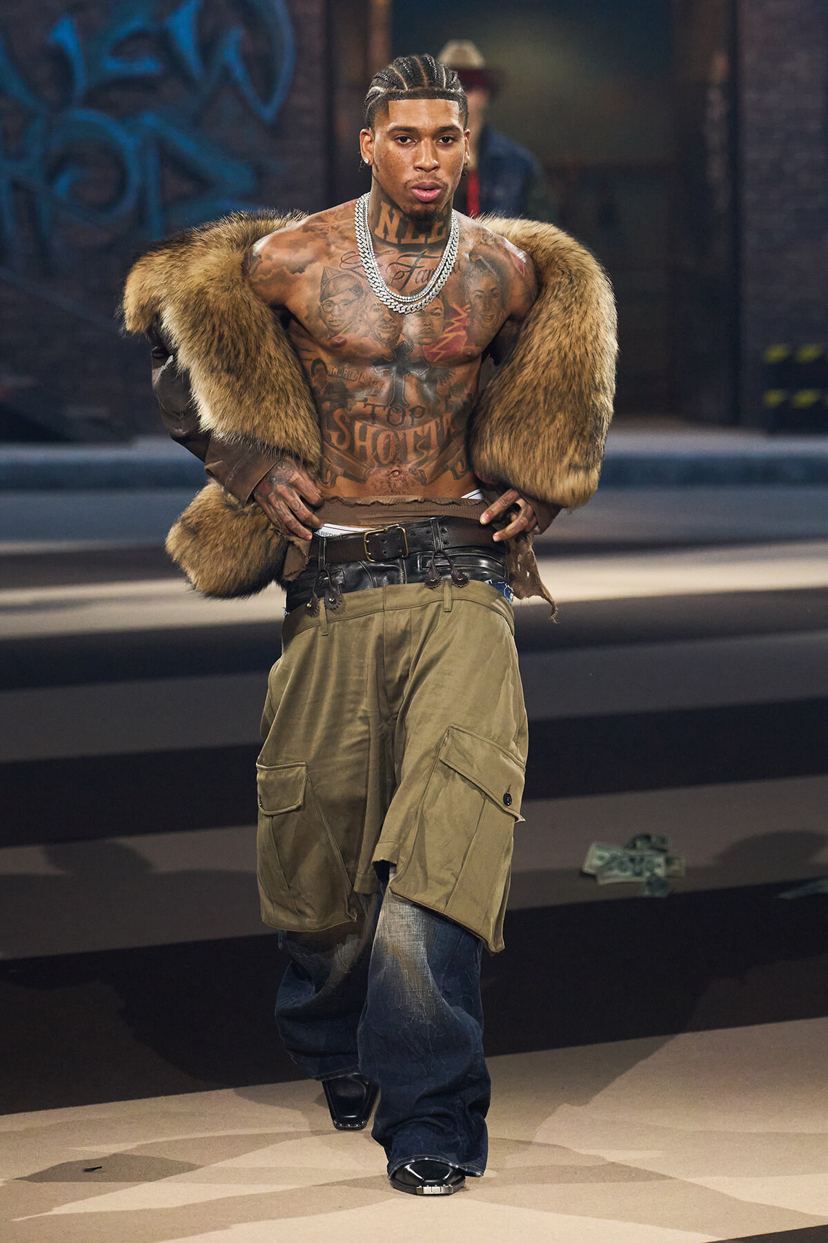 Tattooed male model wearing a fur coat draped over his shoulders, cargo shorts, and jeans, posing in an urban setting.