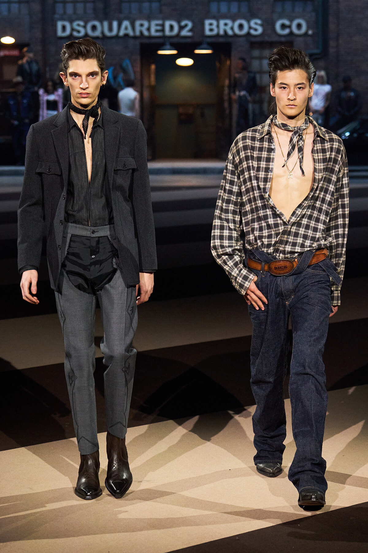 Two male models walk a Dsquared2 runway in Western-inspired outfits: left in a black blazer and sheer shirt with gray trousers, right in a plaid shirt, jeans, and a leather belt. Behind them, the sign reads DSQUARED2 BROS CO.