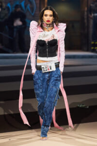 Model walking a fashion runway in a pale pink oversized coat, black corset top over a white graphic tee, blue cargo jeans, and a belt with a New York license plate; long pink scarf trails behind her.