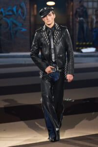 Male model walks a runway in a black studded leather biker jacket, cap, and dark jeans.
