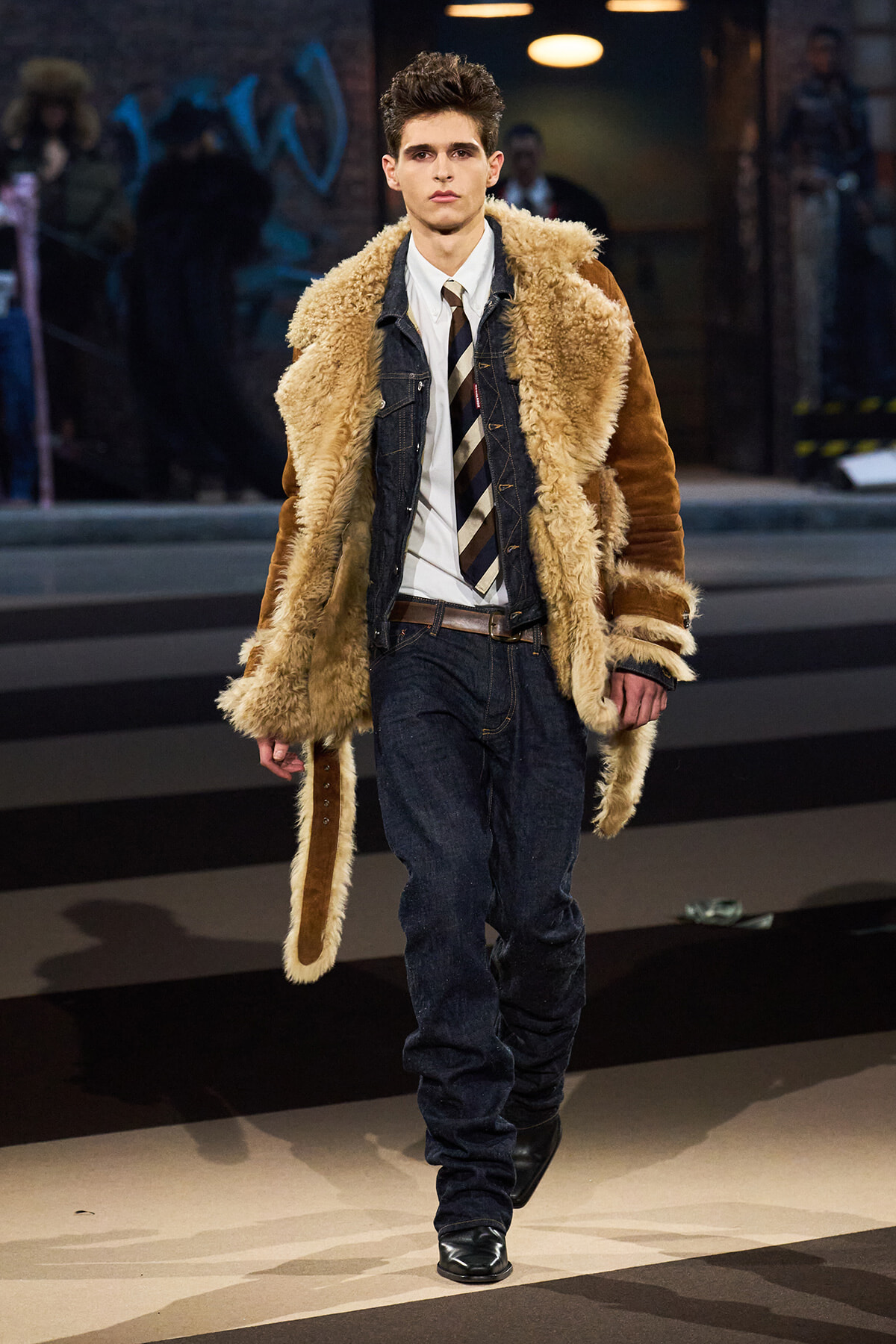 Male model walks the runway in a tan shearling coat over a denim jacket, white shirt, and striped tie.