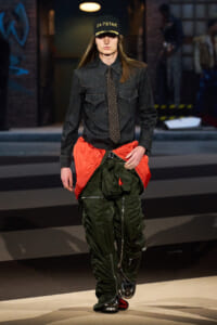 Model walking runway in dark denim shirt, patterned tie, and oversized green cargo pants with bright orange waist panel; black cap reads '24-7 STAR'