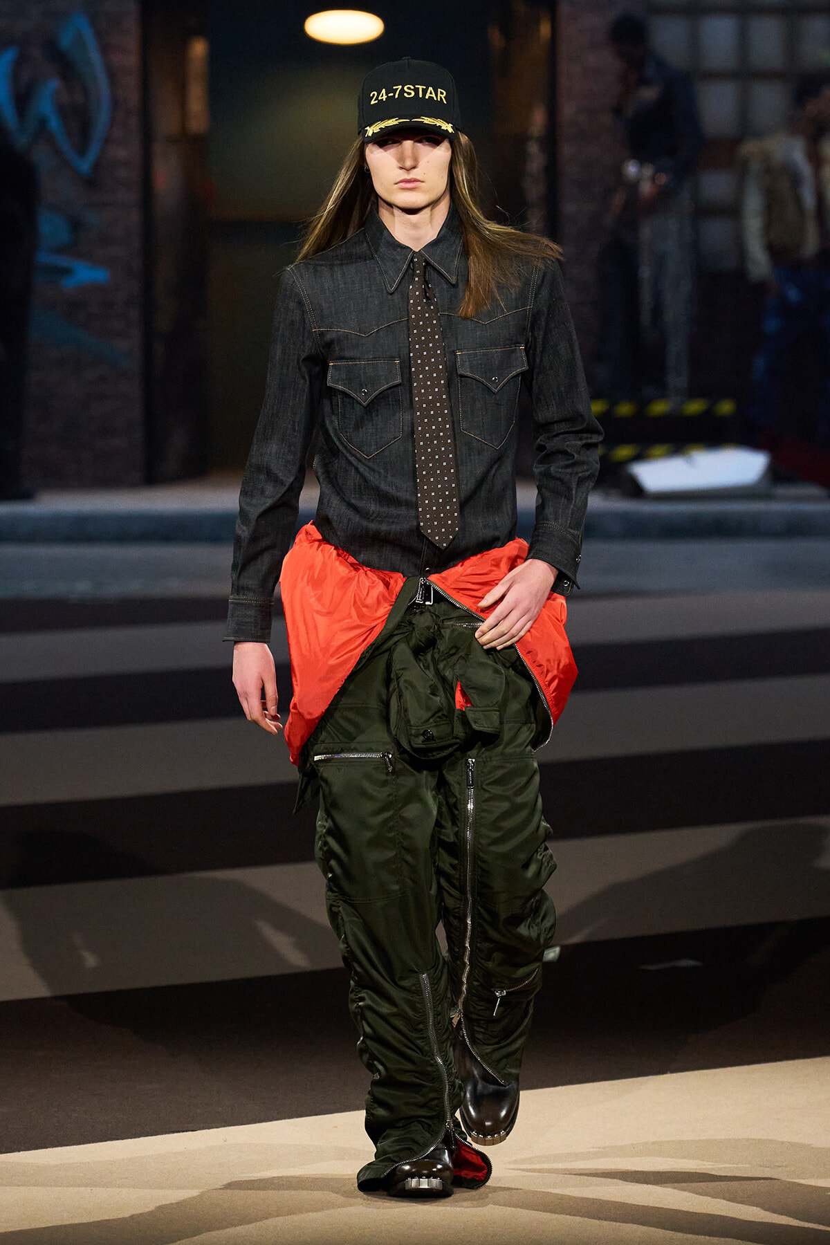 Model walking runway in dark denim shirt, patterned tie, and oversized green cargo pants with bright orange waist panel; black cap reads '24-7 STAR'