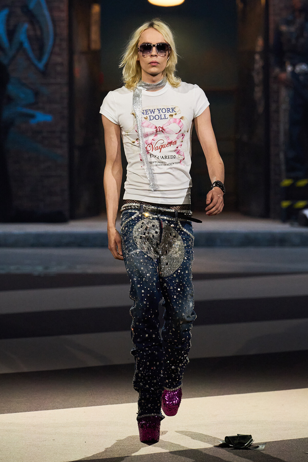 Runway model wearing a white graphic tee, silver scarf, studded embellished jeans, and bright pink glitter boots with sunglasses.