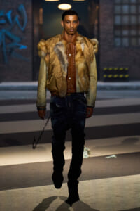 Male model walking on a city street in a tan fur-trimmed bomber jacket over a brown leather shirt with distressed jeans and a hanging chain accessory at his side.