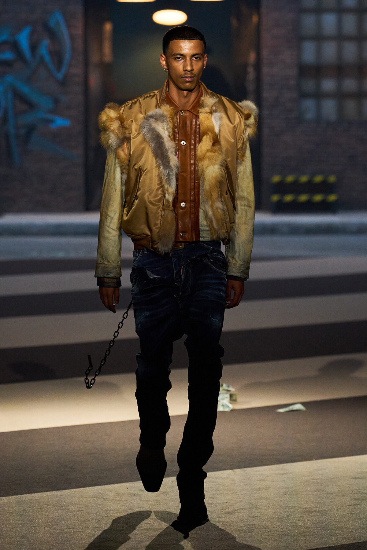 Male model walking on a city street in a tan fur-trimmed bomber jacket over a brown leather shirt with distressed jeans and a hanging chain accessory at his side.