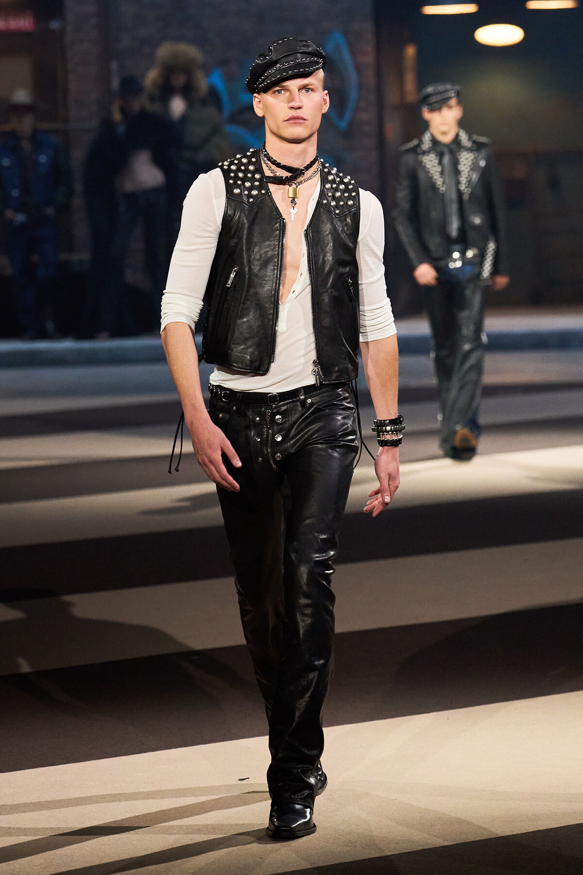 Male model walks a fashion runway in a studded black leather vest over a white long-sleeve shirt, black leather pants, and a cap showcased on the catwalk.