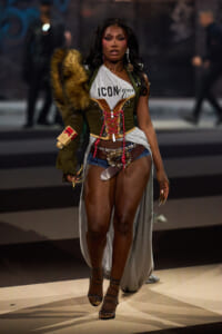 Model walking a fashion show in a corseted olive jacket with fur shoulder, gray cutaway top reading ICONique, denim shorts, and strappy heels.