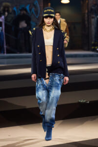 Model walking a runway in a navy double-breasted blazer with gold buttons, paint-splattered baggy jeans, a white bralette, and a black cap that reads '24-7 STAR'.