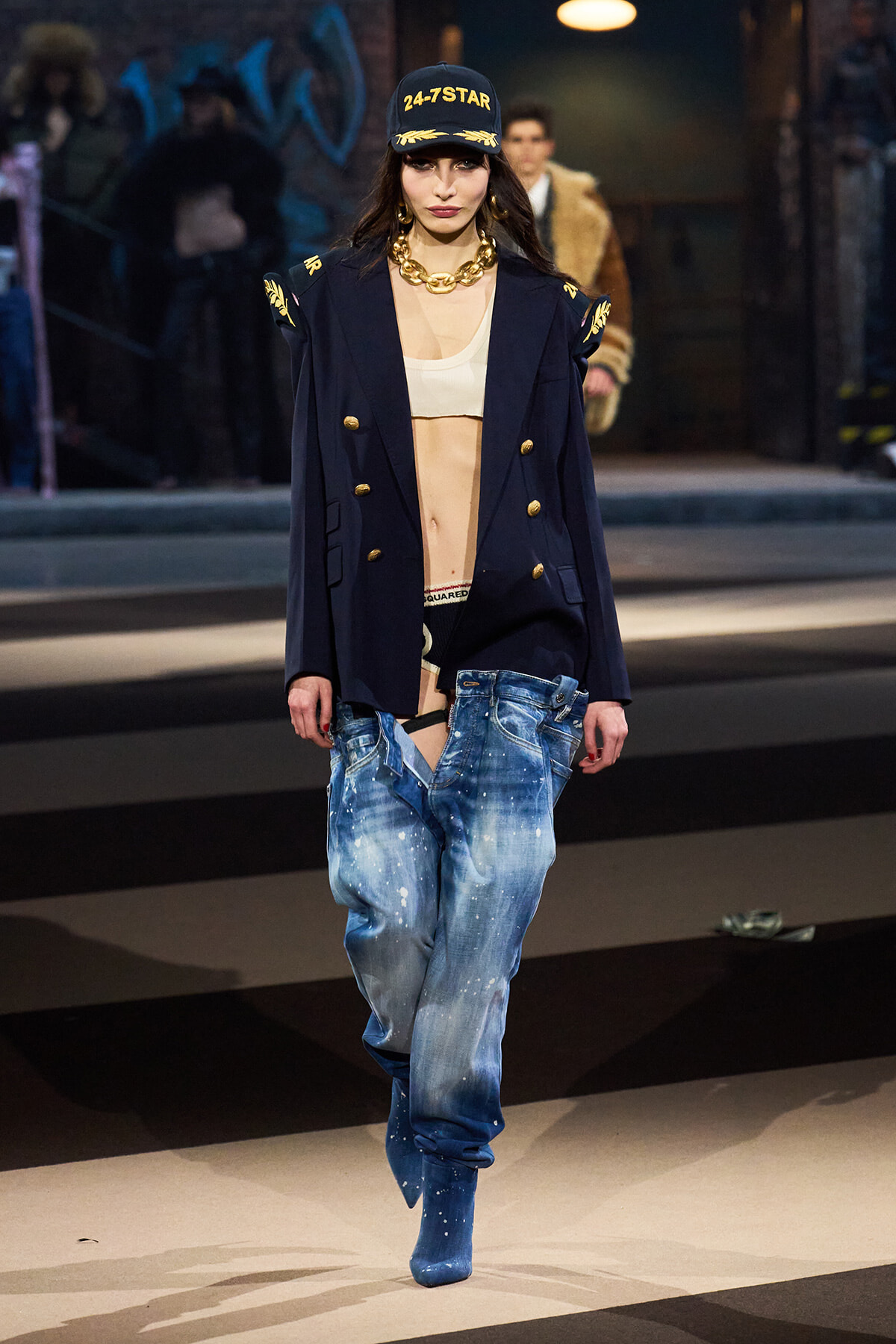 Model walking a runway in a navy double-breasted blazer with gold buttons, paint-splattered baggy jeans, a white bralette, and a black cap that reads '24-7 STAR'.