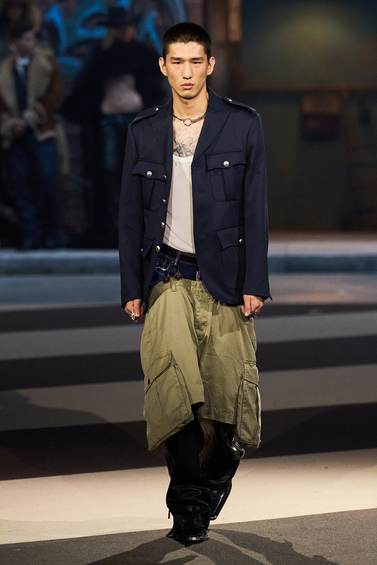 Male model walking a fashion runway in a navy utility jacket over a white tank, with olive cargo shorts and tall black boots; visible chest tattoo and a chain necklace.