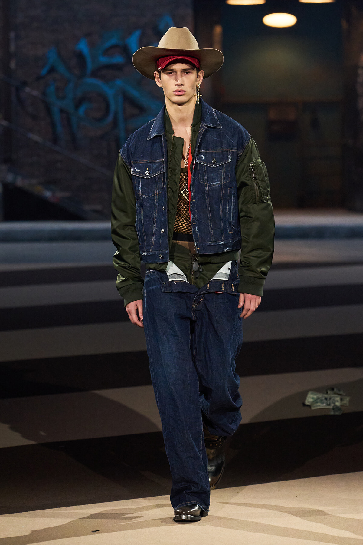 Male model walking a fashion show wearing a denim jacket with olive-green sleeves, baggy jeans, a red bandana, and a tan cowboy hat.
