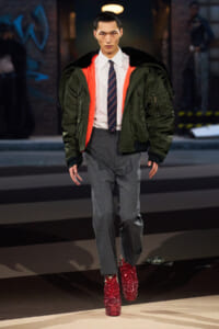 Male model on a runway wearing a green bomber jacket with orange lining over a white shirt, striped tie, gray trousers, and red glitter boots.
