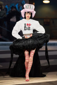 Model on a runway wearing a white cropped hoodie with a heart graphic, an oversized pink hat, a dramatic black skirt, and red lace-up sandals.