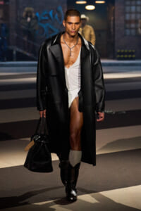 Male model on a runway in a long black coat over a white bodysuit, knee-high socks, black boots, carrying a black bag.