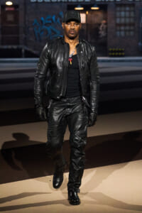 Man in a black leather jacket, pants, gloves, and cap walks toward the camera on a city street with graffiti in the background.