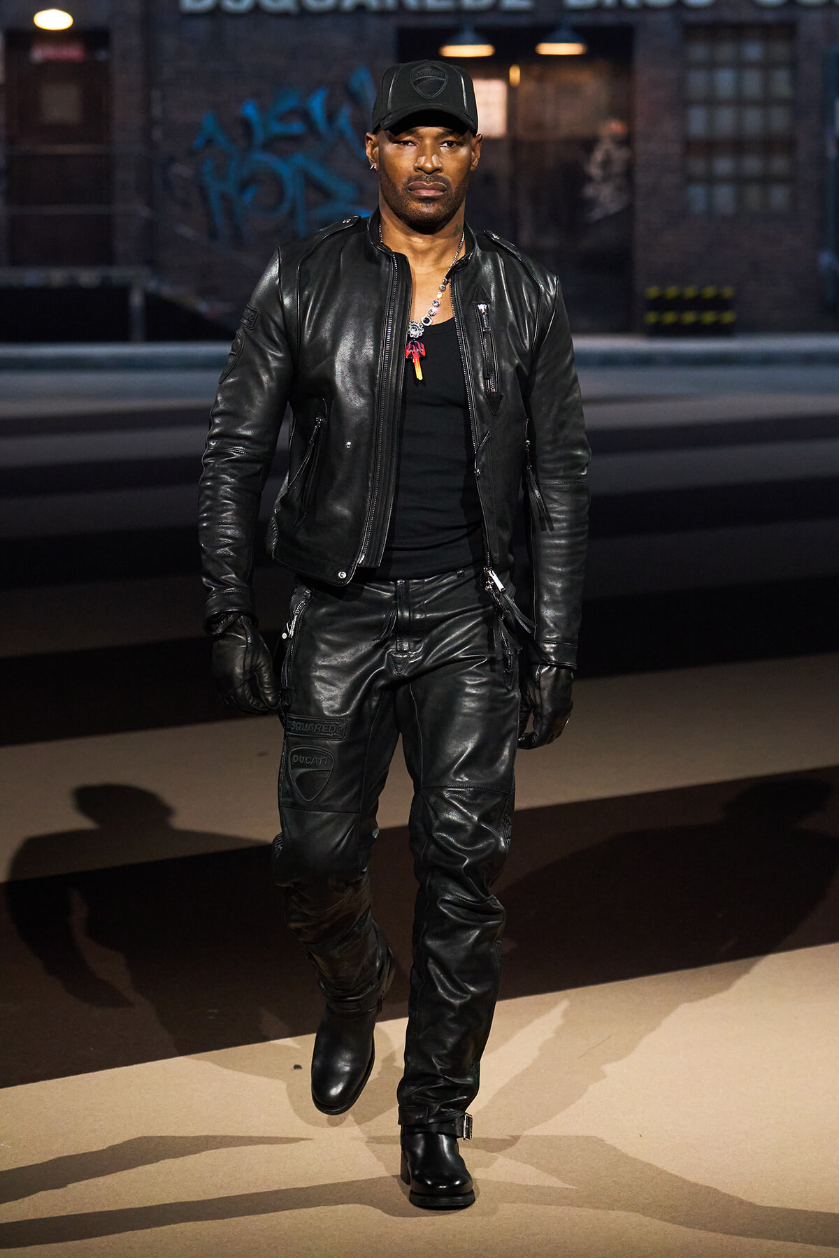 Man in a black leather jacket, pants, gloves, and cap walks toward the camera on a city street with graffiti in the background.