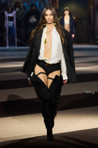 Model struts the runway in an oversized black blazer, white blouse worn open, and dramatic thigh-high garter boots with black shorts beneath.
