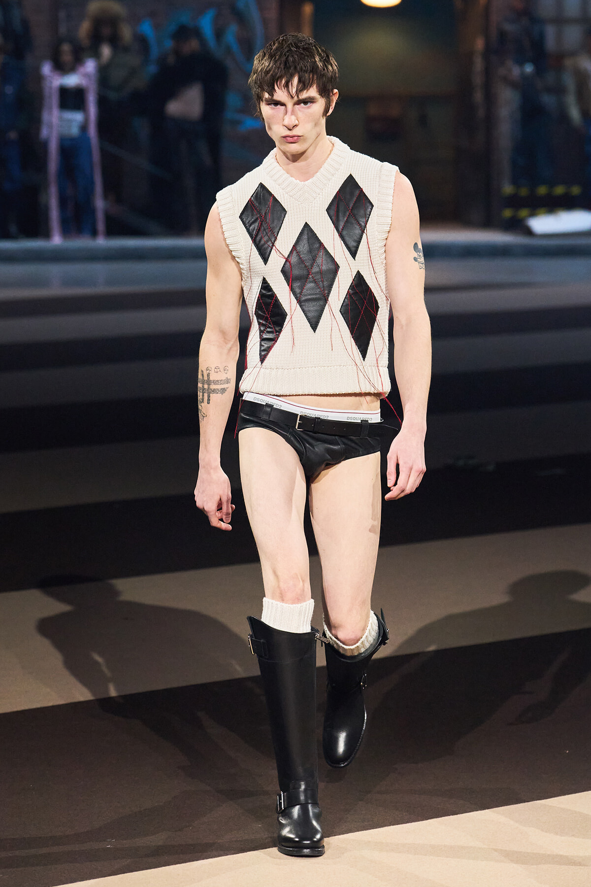 Male model walking a fashion runway in a sleeveless cream argyle-knit sweater, black leather shorts, white socks, and knee-high black boots with visible arm tattoos.
