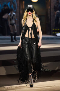 Model walking a runway in a black fringe halter top, leather pants, studded boots, and a cap with a NY logo.