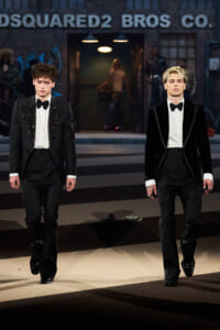 Two male models in black tuxedos walk a runway; one with sequined lapels, the other in a velvet jacket, with a Dsquared2 sign and backstage lighting in the background.