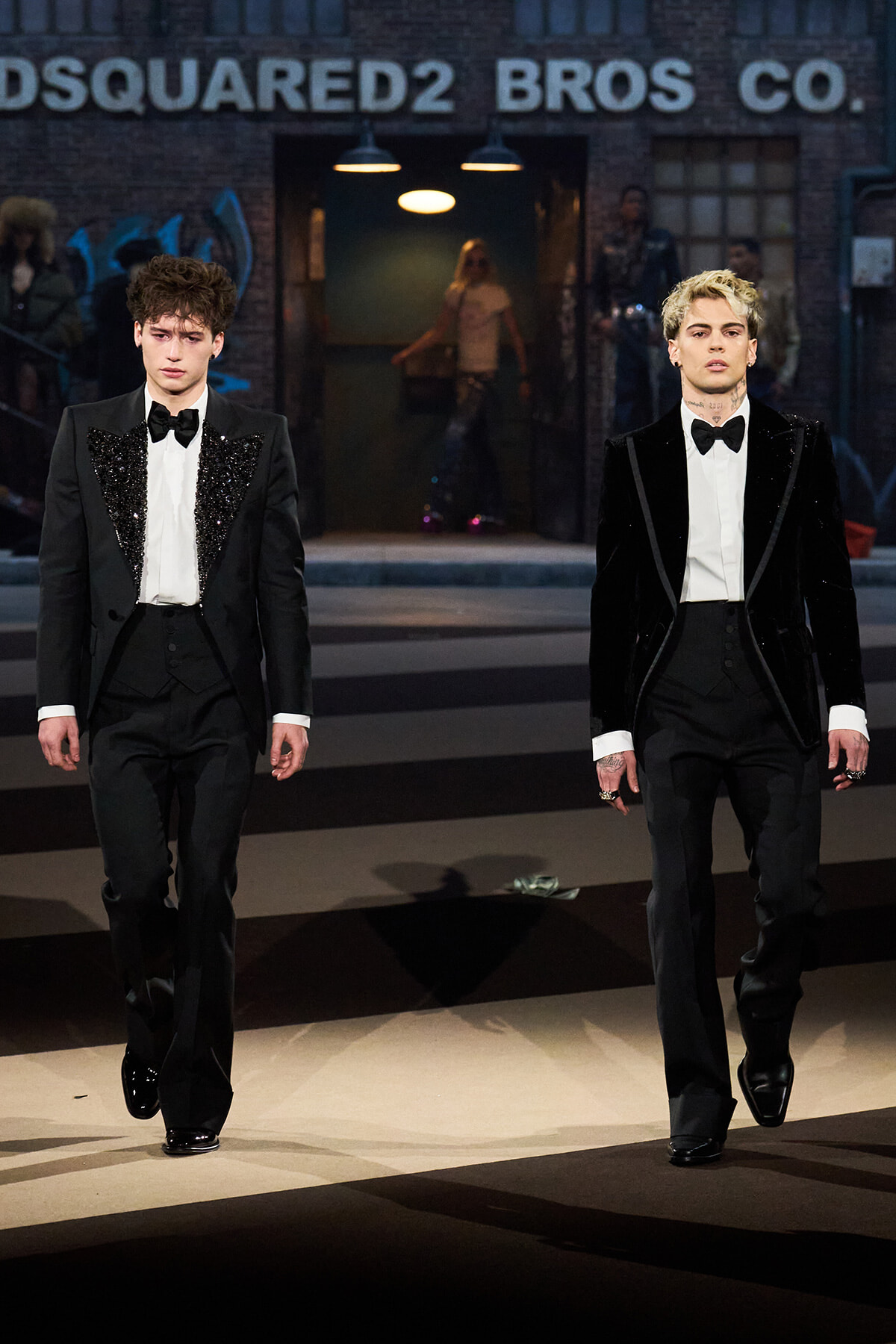 Two male models in black tuxedos walk a runway; one with sequined lapels, the other in a velvet jacket, with a Dsquared2 sign and backstage lighting in the background.