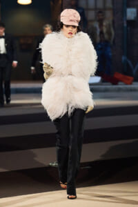 Model on a runway wearing a large white feather coat, pale pink hat, black pants, and open-toe heels, holding a cigarette holder while walking the catwalk.
