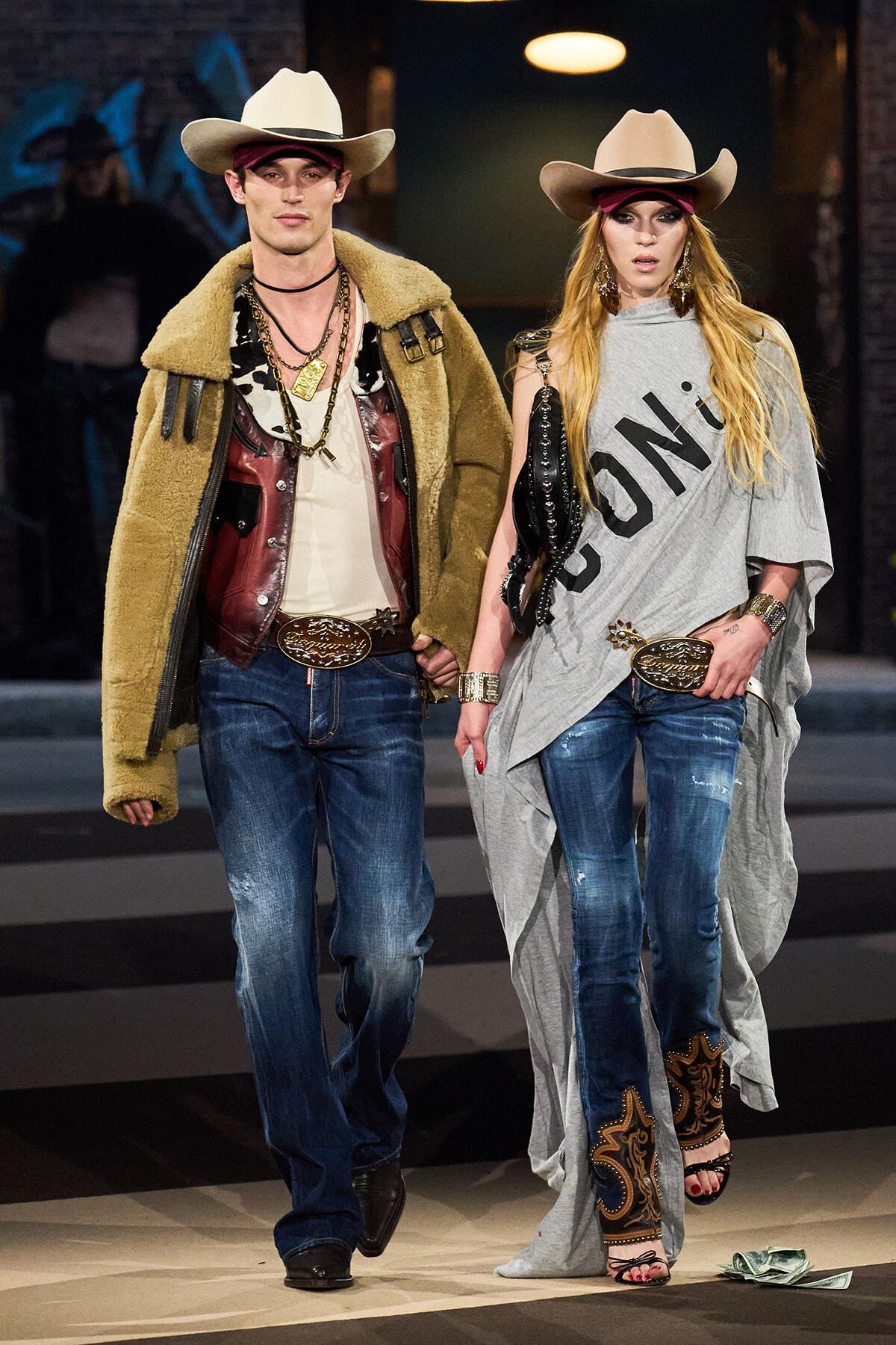 Two models walk a runway in western-inspired outfits: man in tan shearling coat and cowboy hat, woman in gray poncho with lettering, jeans and boots.