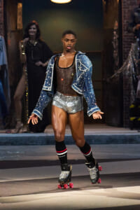 Model roller-skating down a fashion show runway in a studded denim jacket, brown mesh top, and silver shorts, with pink-wheel skates and striped socks.