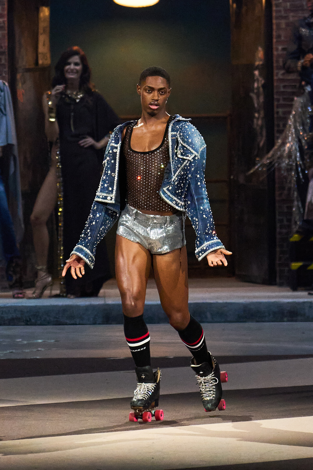 Model roller-skating down a fashion show runway in a studded denim jacket, brown mesh top, and silver shorts, with pink-wheel skates and striped socks.