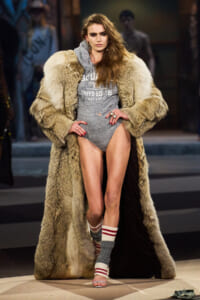 Runway moment: model wearing a gray graphic bodysuit under a long beige fur coat, with striped socks.